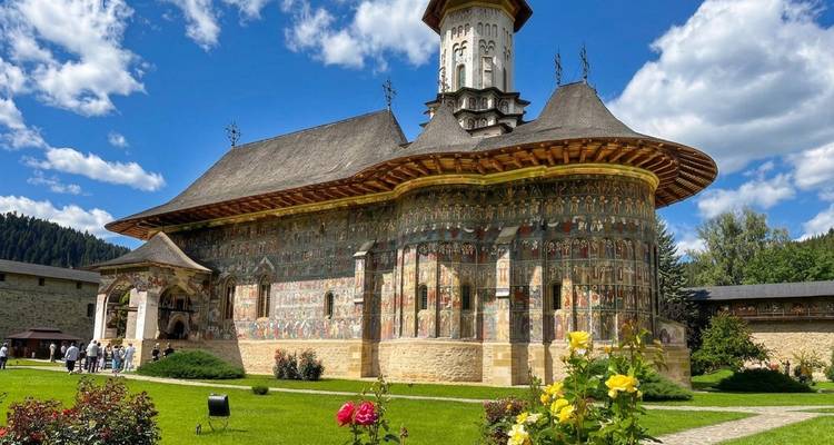 Iglesia medieval bellamente pintada con techo de tejas situada entre jardines bajo un cielo azul brillante y nubes esponjosas