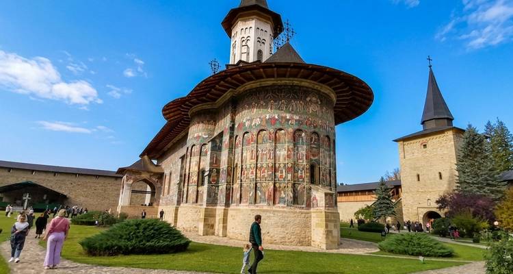 Monasterio de Voronet con frescos exteriores vívidos, torre cónica y visitantes paseando por el patio empedrado bajo cielos azules.