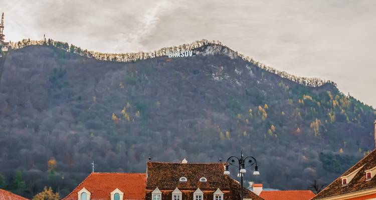 Tejados de un pueblo medieval bajo una ladera montañosa boscosa con el letrero blanco 'BRASOV' en la cima bajo un cielo nublado