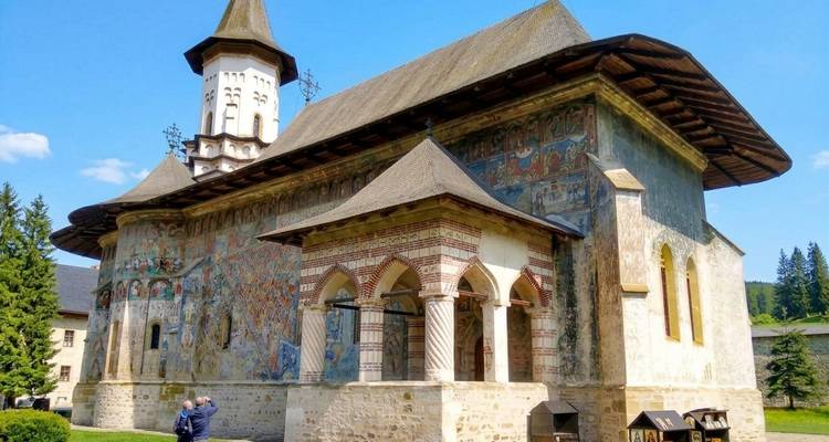 Monasterio rumano ornamentalmente pintado con paredes con frescos, torre alta y visitantes en sus jardines cuidados.