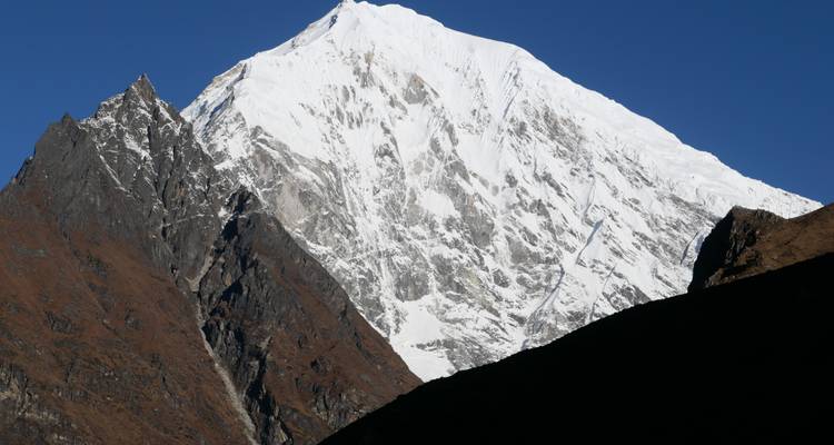 Ein dramatischer, schneebedeckter Gipfel erhebt sich gegen einen tiefblauen Himmel, wobei dunkle Felsgrate seine Seiten rahmen.