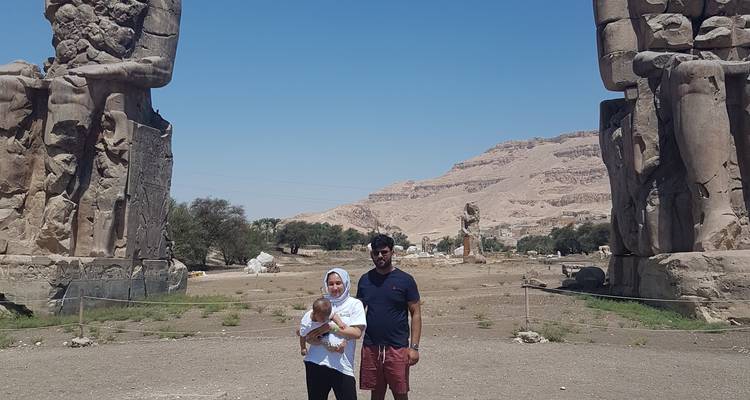 Twee mensen die een kind vasthouden, staand voor grote oude beelden met een woestijnlandschap op de achtergrond.