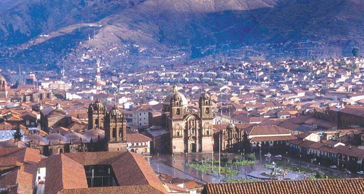 Vista lejana sobre los tejados de barro cocido de Cusco hacia la catedral histórica y las colinas andinas circundantes bajo una luz brumosa.
