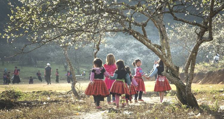 Des filles en robes traditionnelles colorées marchent main dans la main sous des arbres en fleurs tandis que des enfants jouent dans un pré au-delà.