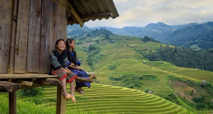 Deux filles souriantes en tenue traditionnelle sont assises sur le rebord d'une maison sur pilotis en bois au-dessus de rizières en terrasses étagées