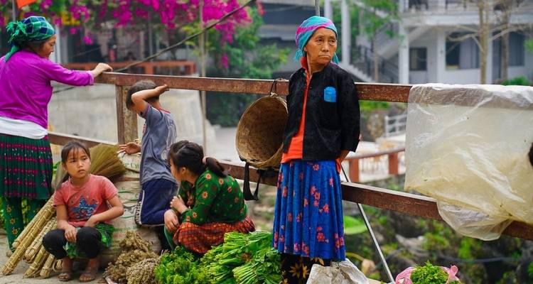 Des villageois, y compris des femmes en jupes à motifs, offrent des produits frais sur un pont rustique.