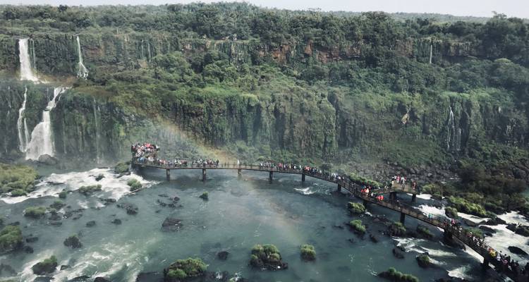 A boardwalk with people above a river with waterfalls and a rainbow.