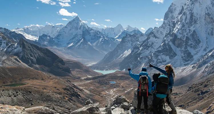 Tres trekkers señalando hacia el otro lado de un enorme valle glacial hacia lagos turquesas y picos del Himalaya.
