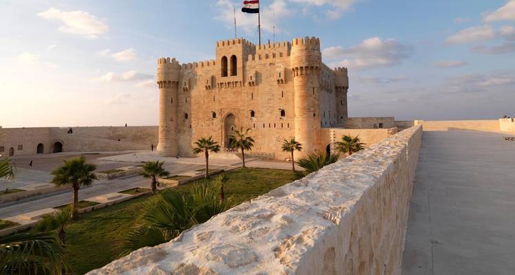 Una fortaleza medieval de piedra con una bandera en la cima, rodeada de palmeras.