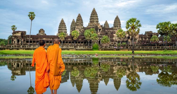 Dos monjes en túnicas naranjas caminando cerca de Angkor Wat con su reflejo visible.
