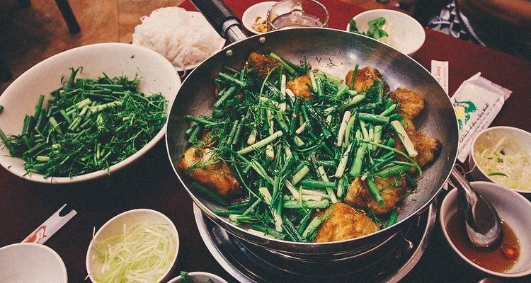 Close-up of a Vietnamese dish with green herbs in a pan surrounded by various side dishes.
