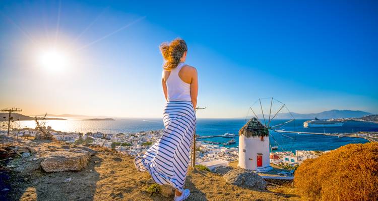 Vrouw in een gestreepte jurk die kijkt naar een zonsondergang zeegezicht met traditionele Griekse windmolen.