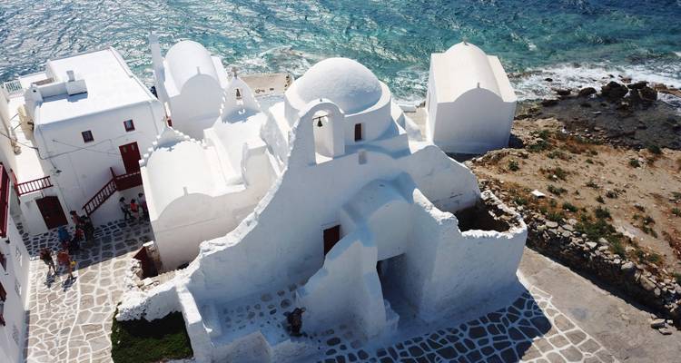 Aerial view of the bright white Paraportiani Church complex on the rocky Mykonos shoreline beside turquoise sea