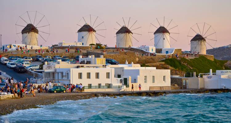 Iconic row of Mykonos windmills overlooking the sea at dusk with crowds along the shoreline