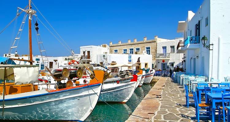 Coloridos barcos de pesca amarrados a lo largo de un muelle de adoquines junto a edificios cicládicos blancos y mesas azules