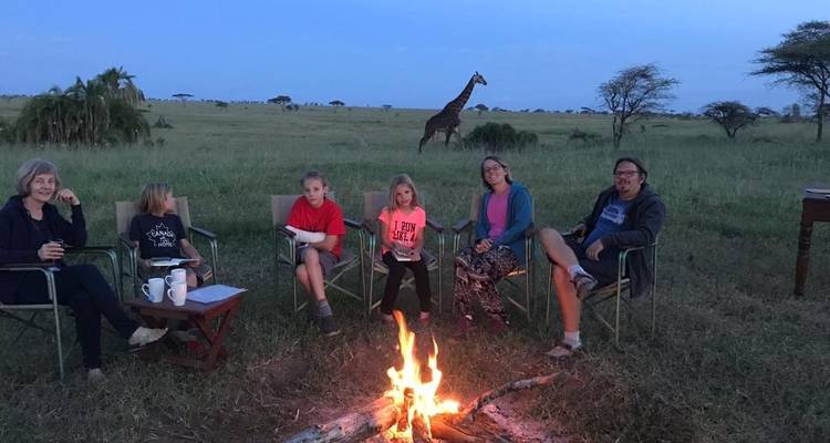 Family sitting around a campfire in the savannah with a giraffe in the background.