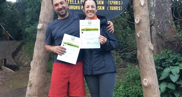 Two people holding certificates in front of a wooden sign in a forest setting.