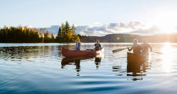Twee rode kano's met glimlachende peddelers glijden over een kalm meer omringd door herfstbos in warm avondlicht.