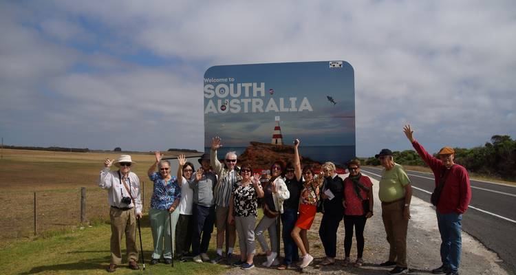 Groupe de touristes posant et saluant à côté d'un panneau routier « Bienvenue en Australie-Méridionale » dans une campagne agricole ouverte.