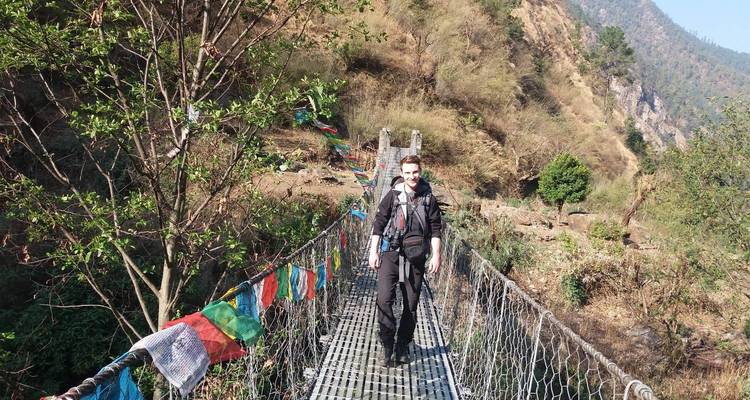 Man die loopt op een hangbrug met gebedsvlaggen in een heuvelachtig gebied.