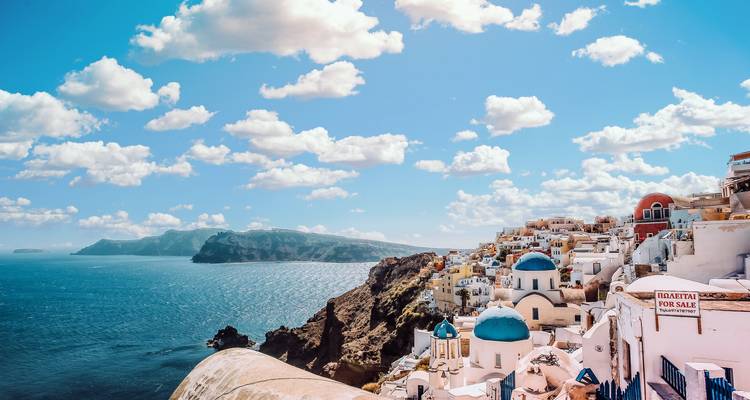 Pintoresco pueblo blanco como la nieve en un acantilado con iglesias de cúpulas azules que se asoman a la caldera y al Mar Egeo bajo un cielo brillante lleno de nubes esponjosas.