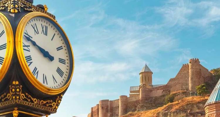 Reloj callejero ornamentado en primer plano con la Fortaleza de Narikala e iglesia en la colina detrás, Tbilisi