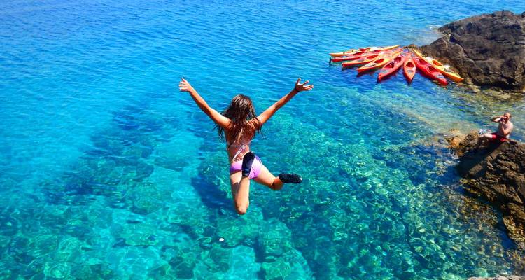 Persona en el aire saltando desde un acantilado hacia el mar Adriático turquesa cristalino cerca de kayaks coloridos