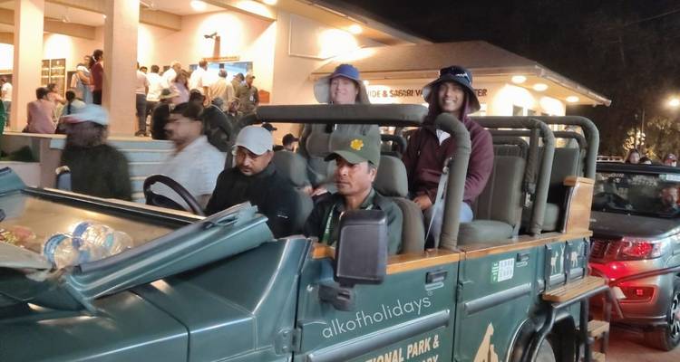 Tourists seated in a dark green safari jeep waiting outside a busy park gate at dusk.