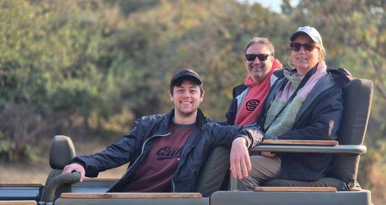 Three smiling tourists sit in an open-air safari vehicle with forest scenery in the background.