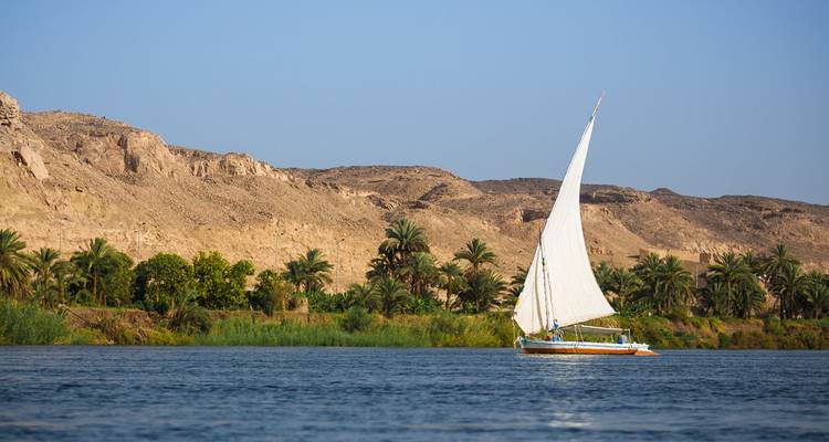 Traditionele felucca met wit zeil glijdt langs de Nijl omgeven door palmen en droge heuvels.