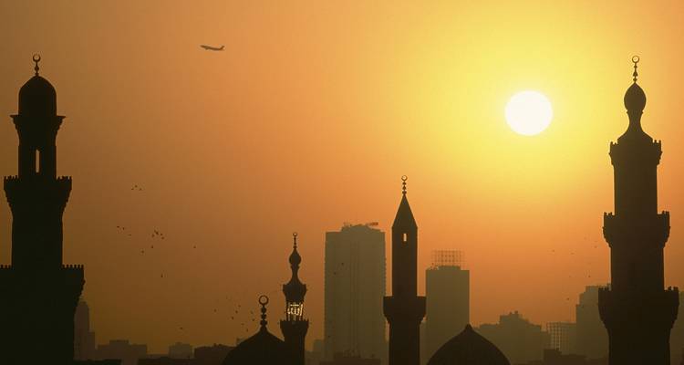 Silhouette de minarets et de bâtiments au coucher du soleil avec un avion dans le ciel.
