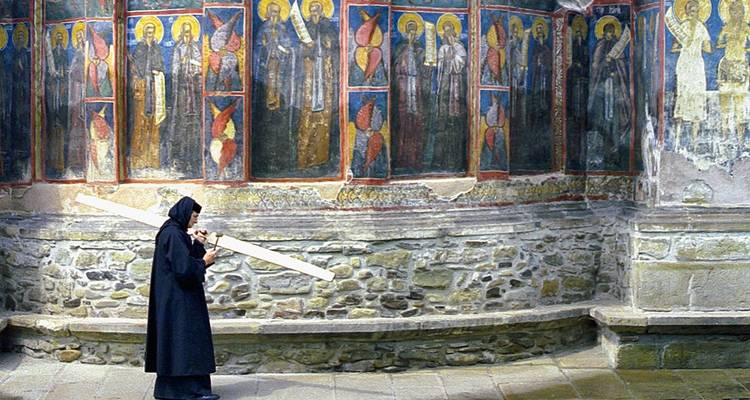A robed monk carrying a wooden beam walks along a vividly painted fresco wall inside a stone monastery corridor.