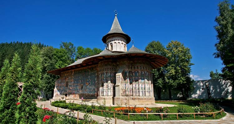 A beautifully painted Orthodox monastery with conical roof set amid gardens under a clear blue sky.