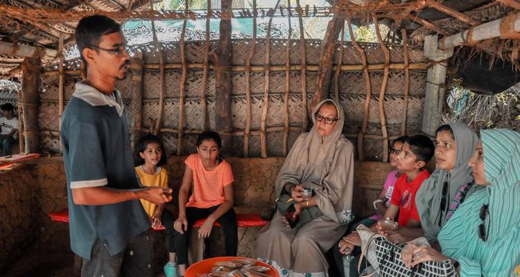 Group of people sitting in a rustic hut, listening to a man speak.