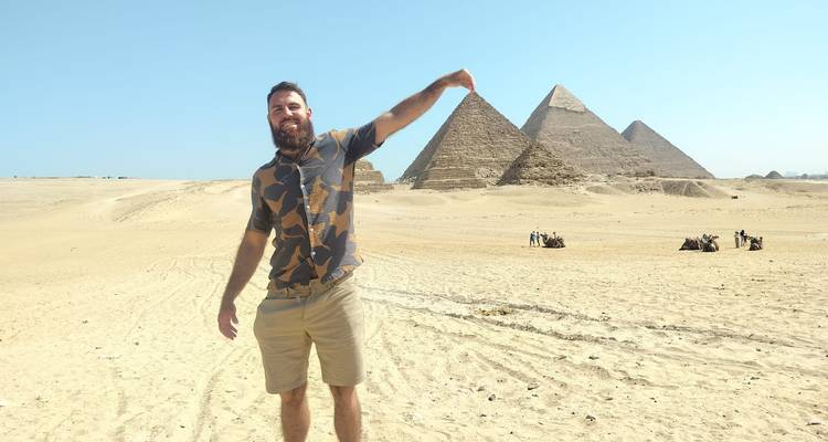 Mann, der vor den Pyramiden von Gizeh posiert und so tut, als würde er die Spitze einer Pyramide zwischen den Fingern halten.