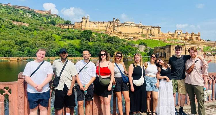 Lächelnde Reisegruppe posiert auf einer Brücke mit den massiven gelben Mauern des Amber Fort, die sich auf einem üppig grünen Hügel hinter ihnen erheben