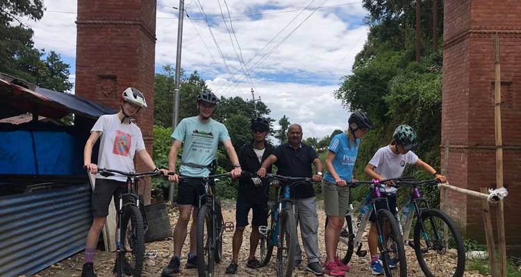 A small cycling group poses with mountain bikes near a brick gate under patchy clouds.