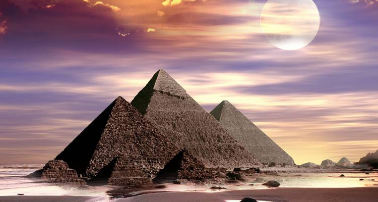 Vista dramática de las tres pirámides de Giza elevándose desde el desierto contra un cielo al atardecer lleno de colores con una luna grande arriba y suaves olas rompiendo en la arenosa zona delantera.