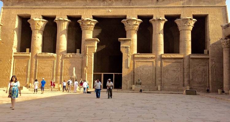 Los visitantes exploran la gran entrada de arenisca y las columnas talladas de un antiguo templo egipcio bañado por la cálida luz del sol.