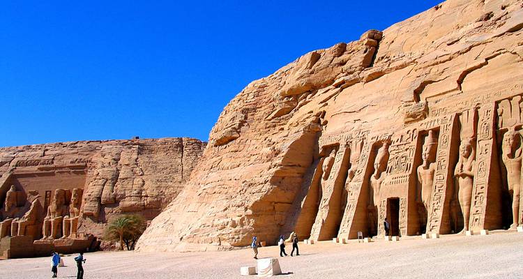 Vista expansiva de los templos excavados en roca de Abu Simbel contra un cielo azul vivido, con pequeños turistas que dan una idea de la escala.