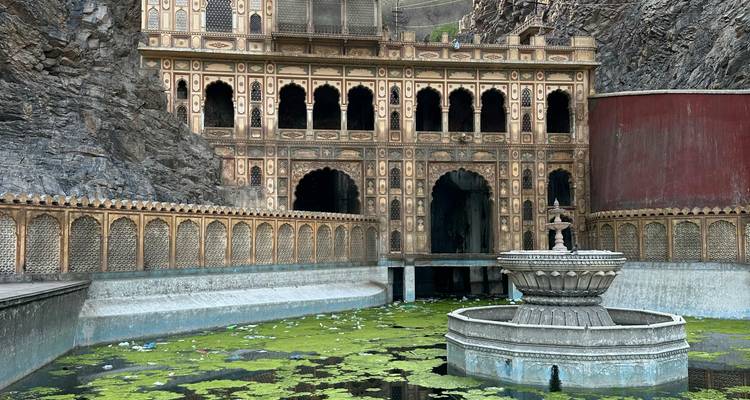 Sierlijke zandstenen gevel van de Galtaji-tempel tegen rotsachtige kliffen met een decoratieve vijver en fontein.