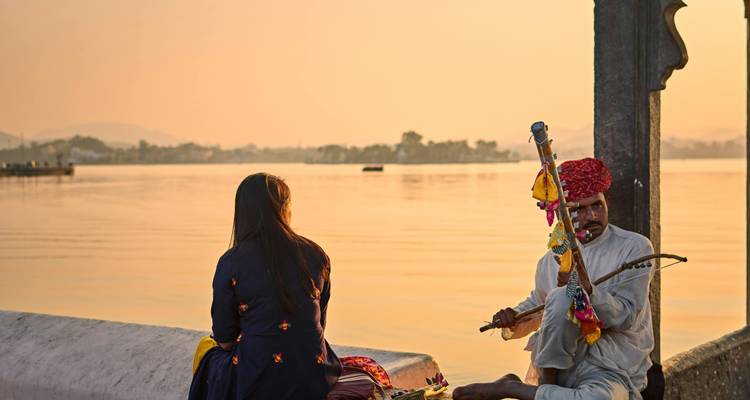 Traditionele Rajasthani-muzikant die speelt bij een serene meer bij zonsondergang terwijl een vrouw over het water kijkt.