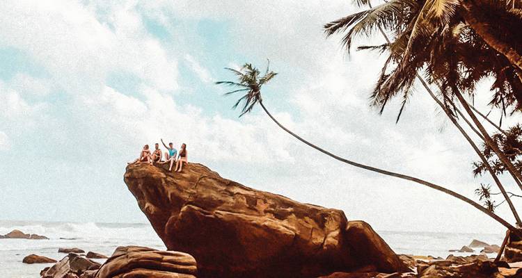Friends perch atop a dramatic coastal boulder beside leaning palm trees and crashing tropical surf.