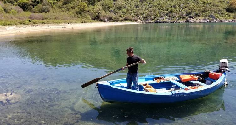 Hombre remando un bote azul en un lago cristalino con un bosque frondoso de fondo.