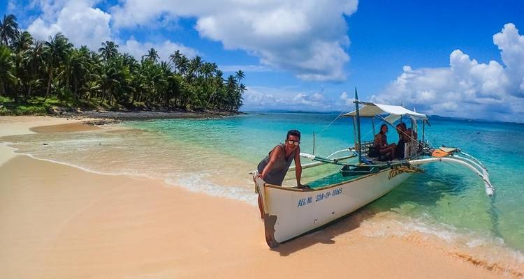 Menschen auf einem Boot an einem tropischen Strand mit blauem Himmel und Palmen.