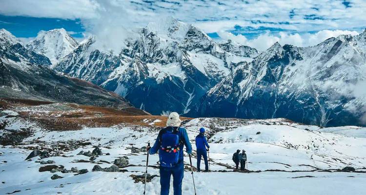 Trekkingstokers die over een besneeuwde bergrug lopen met dramatische Himalaya-pieken die uitsteken tegen een gedeeltelijk bewolkte hemel.