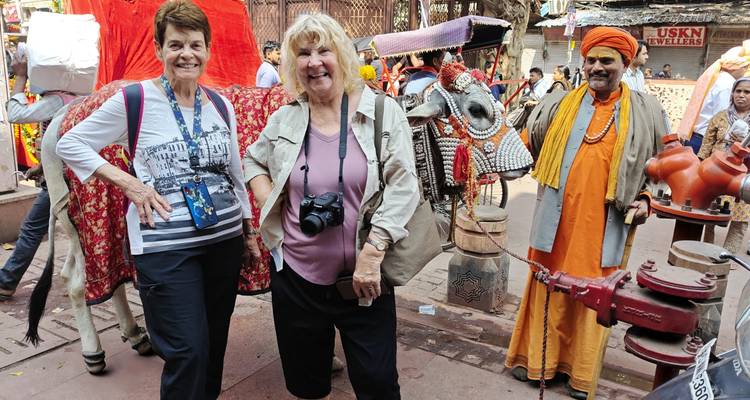 Dos mujeres viajeras posando con cámaras junto a un carrito de bueyes decorado y un sadhu con ropas de colores brillantes en una bulliciosa calle del mercado indio.