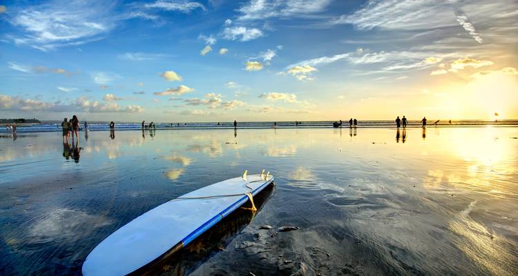 Surfboard on reflective wet sand at sunset with people