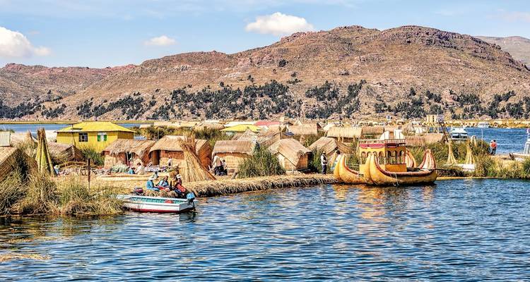 Las islas de totora y una colorida barca de totora flotan en las aguas azul brillante del lago Titicaca con colinas áridas de fondo.
