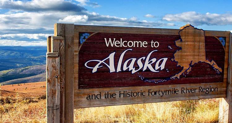 Panneau routier en bois indiquant « Bienvenue en Alaska » avec des collines ondulantes au loin et des nuages spectaculaires en arrière-plan.